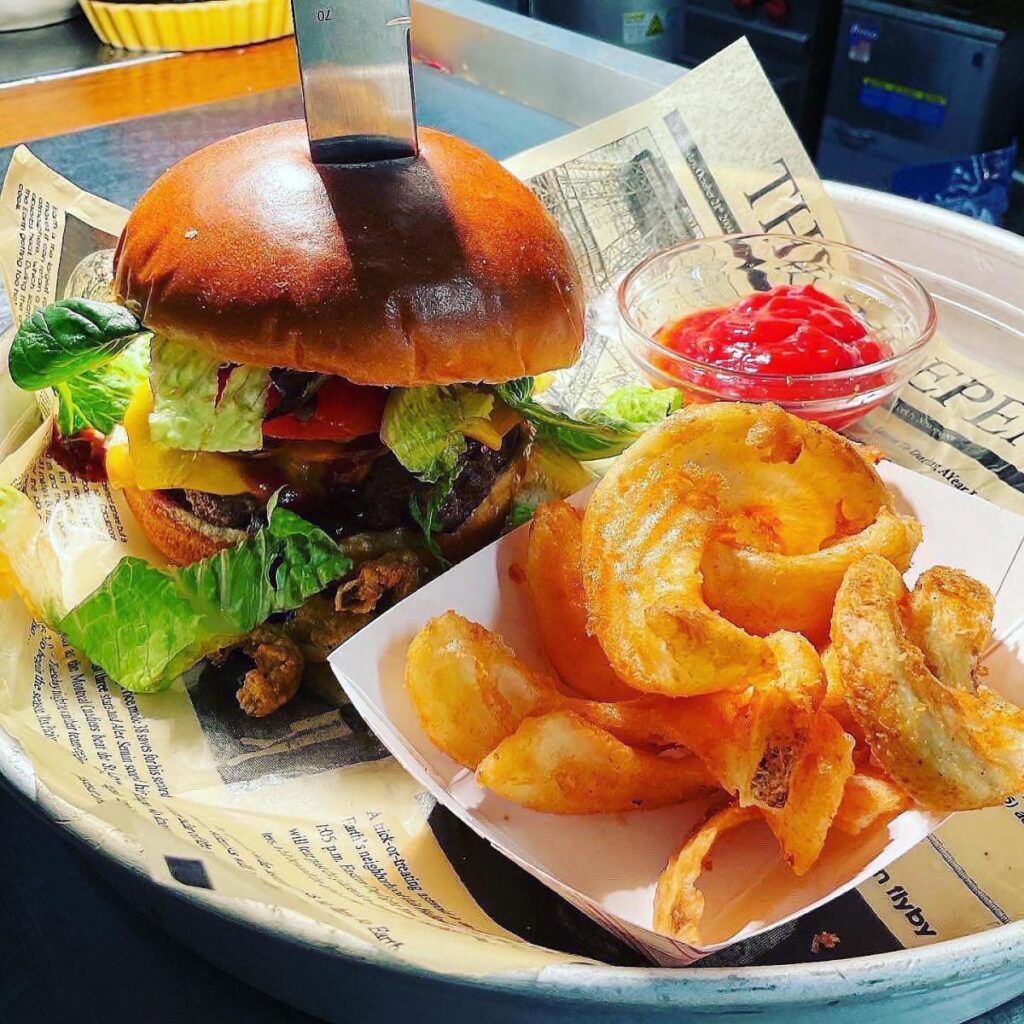 A cheeseburger piled high with toppings and a side of French fries served on a white plate lined with newspaper