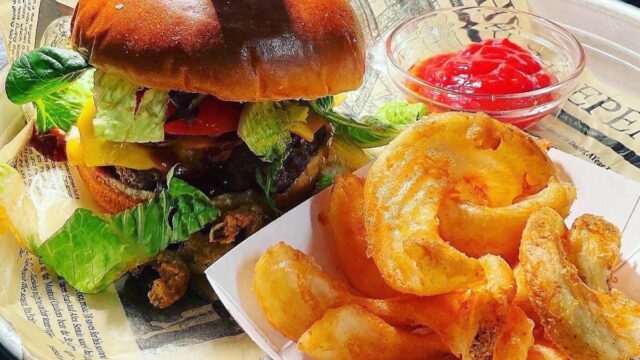 A cheeseburger piled high with toppings and a side of French fries served on a white plate lined with newspaper