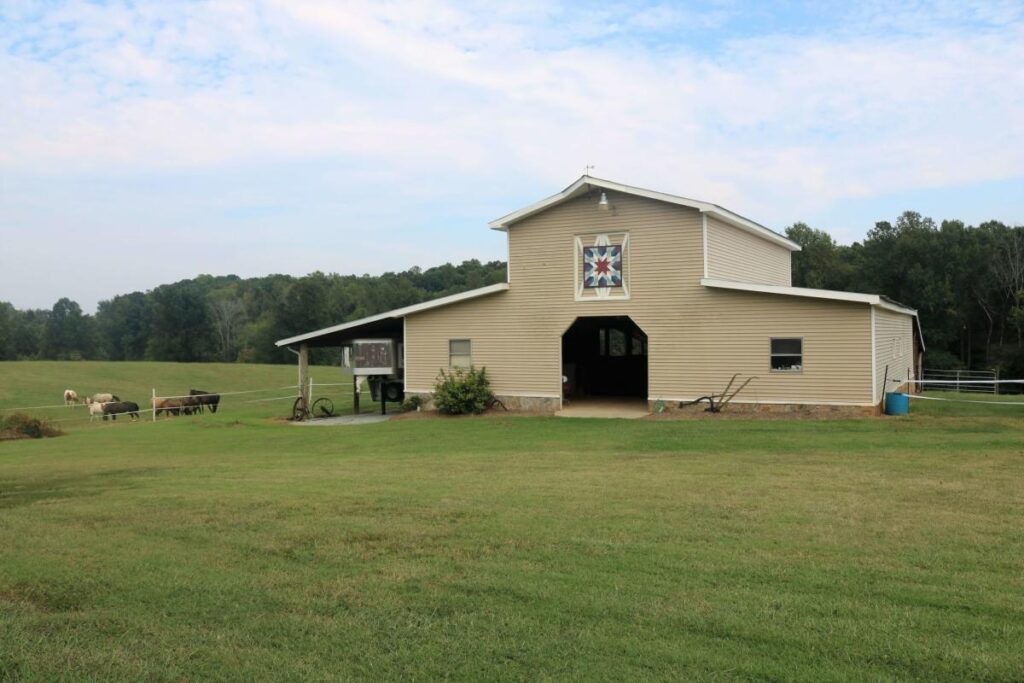 A tan barn surrounded by green grass has a painted quilt above the entrance