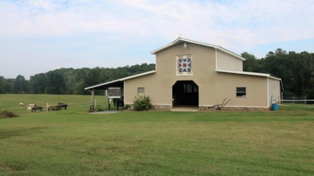A tan barn surrounded by green grass has a painted quilt above the entrance