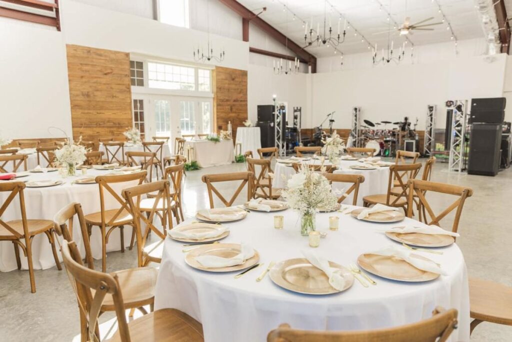 Round tables covered with white tablecloths and set with dishes, cutlery, and folded napkins near a dance floor space with instruments and speakers