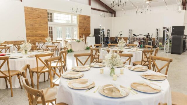 Round tables covered with white tablecloths and set with dishes, cutlery, and folded napkins near a dance floor space with instruments and speakers